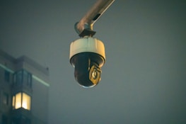 A surveillance camera is mounted on an extended arm against a background of a building at night. The building has a few lit windows, and the overall scene is dimly lit.