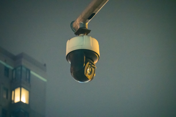 A surveillance camera is mounted on an extended arm against a background of a building at night. The building has a few lit windows, and the overall scene is dimly lit.