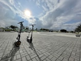 Two electric scooters are parked on a wide paved area under a partly cloudy sky with the sun visible. The background includes trees, benches, and distant buildings, creating a spacious and open urban environment.