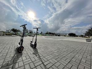 Two electric scooters are parked on a wide paved area under a partly cloudy sky with the sun visible. The background includes trees, benches, and distant buildings, creating a spacious and open urban environment.