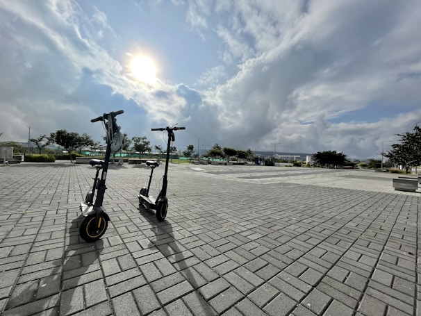 Two electric scooters are parked on a wide paved area under a partly cloudy sky with the sun visible. The background includes trees, benches, and distant buildings, creating a spacious and open urban environment.