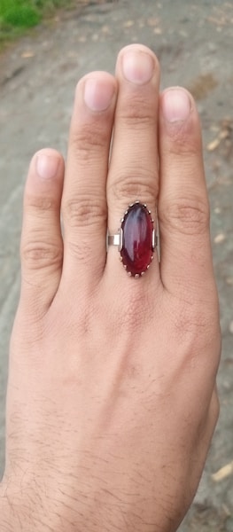 A hand displaying a silver ring with an oval red gemstone, set against an outdoor backdrop of pavement and some green foliage.