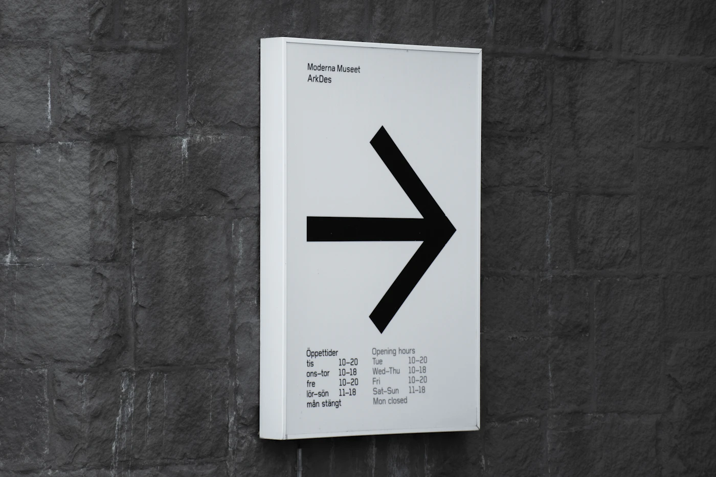 A white directional wayfinding sign with a bold arrow and bilingual opening hours, mounted on a stone wall