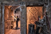 A man stands in a rustic workshop, examining a piece of wood. The space is filled with woodworking tools and materials. To the right, a rooster and a nearby hen perch on large logs, adding a rural, natural element to the scene. The stone walls and archways of the building create an old-world charm.