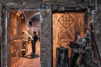 A man stands in a rustic workshop, examining a piece of wood. The space is filled with woodworking tools and materials. To the right, a rooster and a nearby hen perch on large logs, adding a rural, natural element to the scene. The stone walls and archways of the building create an old-world charm.