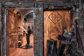 A man stands in a rustic workshop, examining a piece of wood. The space is filled with woodworking tools and materials. To the right, a rooster and a nearby hen perch on large logs, adding a rural, natural element to the scene. The stone walls and archways of the building create an old-world charm.