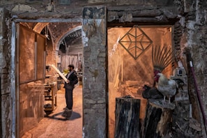 A man stands in a rustic workshop, examining a piece of wood. The space is filled with woodworking tools and materials. To the right, a rooster and a nearby hen perch on large logs, adding a rural, natural element to the scene. The stone walls and archways of the building create an old-world charm.