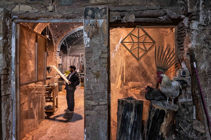 A man stands in a rustic workshop, examining a piece of wood. The space is filled with woodworking tools and materials. To the right, a rooster and a nearby hen perch on large logs, adding a rural, natural element to the scene. The stone walls and archways of the building create an old-world charm.