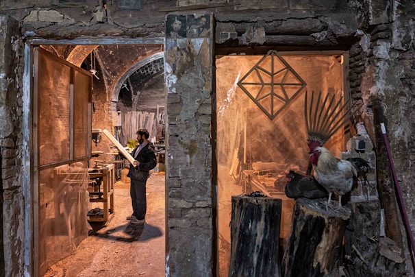 A man stands in a rustic workshop, examining a piece of wood. The space is filled with woodworking tools and materials. To the right, a rooster and a nearby hen perch on large logs, adding a rural, natural element to the scene. The stone walls and archways of the building create an old-world charm.