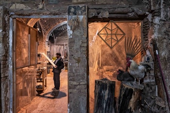 A man stands in a rustic workshop, examining a piece of wood. The space is filled with woodworking tools and materials. To the right, a rooster and a nearby hen perch on large logs, adding a rural, natural element to the scene. The stone walls and archways of the building create an old-world charm.