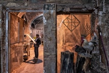 A man stands in a rustic workshop, examining a piece of wood. The space is filled with woodworking tools and materials. To the right, a rooster and a nearby hen perch on large logs, adding a rural, natural element to the scene. The stone walls and archways of the building create an old-world charm.