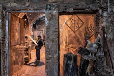 A man stands in a rustic workshop, examining a piece of wood. The space is filled with woodworking tools and materials. To the right, a rooster and a nearby hen perch on large logs, adding a rural, natural element to the scene. The stone walls and archways of the building create an old-world charm.