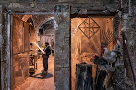 A man stands in a rustic workshop, examining a piece of wood. The space is filled with woodworking tools and materials. To the right, a rooster and a nearby hen perch on large logs, adding a rural, natural element to the scene. The stone walls and archways of the building create an old-world charm.