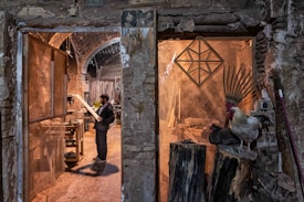 A man stands in a rustic workshop, examining a piece of wood. The space is filled with woodworking tools and materials. To the right, a rooster and a nearby hen perch on large logs, adding a rural, natural element to the scene. The stone walls and archways of the building create an old-world charm.