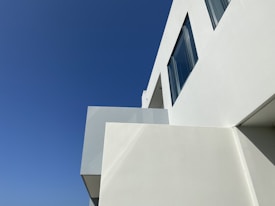A modern, minimalist building with clean lines and large windows set against a clear blue sky. The building is primarily white, emphasizing its sleek architectural design.