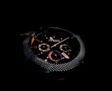 A close-up of a dark, sophisticated wristwatch with a textured metallic bezel and detailed sub-dials on the face. The brand name 'Fossil' is visible, and the design features a combination of polished and matte elements.
