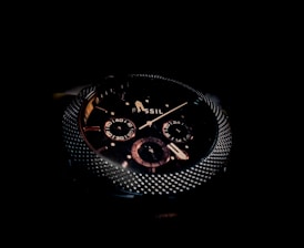 A close-up of a dark, sophisticated wristwatch with a textured metallic bezel and detailed sub-dials on the face. The brand name 'Fossil' is visible, and the design features a combination of polished and matte elements.