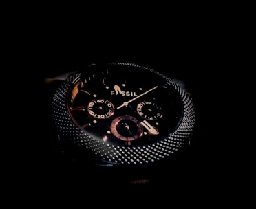 A close-up of a dark, sophisticated wristwatch with a textured metallic bezel and detailed sub-dials on the face. The brand name 'Fossil' is visible, and the design features a combination of polished and matte elements.