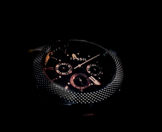 A close-up of a dark, sophisticated wristwatch with a textured metallic bezel and detailed sub-dials on the face. The brand name 'Fossil' is visible, and the design features a combination of polished and matte elements.