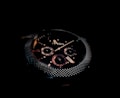 A close-up of a dark, sophisticated wristwatch with a textured metallic bezel and detailed sub-dials on the face. The brand name 'Fossil' is visible, and the design features a combination of polished and matte elements.