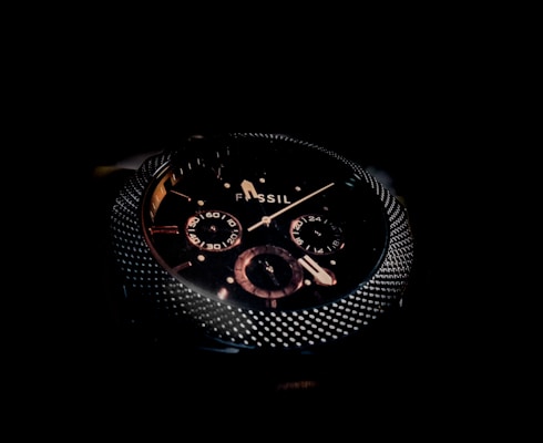 A close-up of a dark, sophisticated wristwatch with a textured metallic bezel and detailed sub-dials on the face. The brand name 'Fossil' is visible, and the design features a combination of polished and matte elements.
