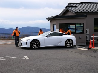 A white sports car is parked near a building with a sloped roof. Two people wearing orange jackets and caps are standing on either side of the car, appearing to be directing it or observing something. The background reveals a scenic view of mountains under a cloudy sky. Bright orange traffic cones are visible near the building.