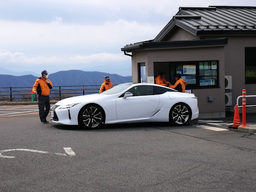 A white sports car is parked near a building with a sloped roof. Two people wearing orange jackets and caps are standing on either side of the car, appearing to be directing it or observing something. The background reveals a scenic view of mountains under a cloudy sky. Bright orange traffic cones are visible near the building.