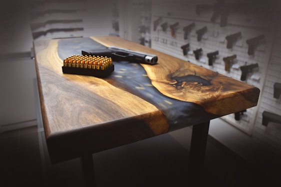 A wooden table with a polished surface, featuring a dark, natural pattern and a matte black finish along the edges. On the table sits a handgun and a box of ammunition. The background includes a wall with various guns displayed.