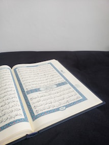 An open book with Arabic script, likely a religious text, resting on a dark surface. The pages display ornate blue and red borders and clear black text. The background is a plain, light-colored wall, creating a calm and focused setting.