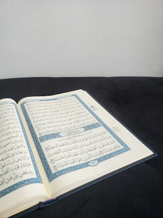 An open book with Arabic script, likely a religious text, resting on a dark surface. The pages display ornate blue and red borders and clear black text. The background is a plain, light-colored wall, creating a calm and focused setting.