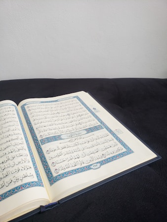 An open book with Arabic script, likely a religious text, resting on a dark surface. The pages display ornate blue and red borders and clear black text. The background is a plain, light-colored wall, creating a calm and focused setting.