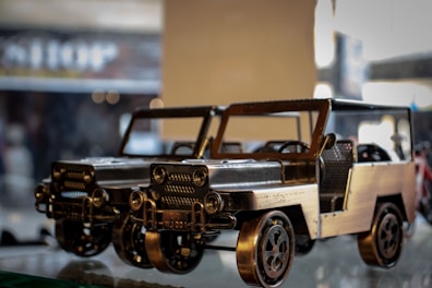 Two metallic model cars with intricate details are displayed on a reflective surface. The design is vintage with an open-roof and visible steering wheels. The background is blurred, giving a focus on the models.