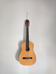 An acoustic guitar is hanging against a plain, light-colored wall. The guitar's body is made of a warm, light brown wood, and it features six strings and a classic design.