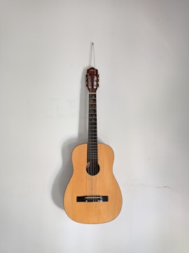 An acoustic guitar is hanging against a plain, light-colored wall. The guitar's body is made of a warm, light brown wood, and it features six strings and a classic design.