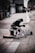 A person dressed in a winter coat and hat sits on a city street, huddled over with a quilt draped over their knees. Next to them is an acoustic guitar lying on the ground, and a cardboard sign that reads 'THANK YOU 4 UR HELP & SUPPORTING THE HOMELESS' is propped up against a container. The setting suggests an urban environment with buildings lining the street.