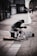 A person dressed in a winter coat and hat sits on a city street, huddled over with a quilt draped over their knees. Next to them is an acoustic guitar lying on the ground, and a cardboard sign that reads 'THANK YOU 4 UR HELP & SUPPORTING THE HOMELESS' is propped up against a container. The setting suggests an urban environment with buildings lining the street.