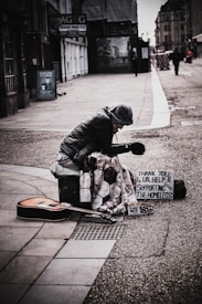 A person dressed in a winter coat and hat sits on a city street, huddled over with a quilt draped over their knees. Next to them is an acoustic guitar lying on the ground, and a cardboard sign that reads 'THANK YOU 4 UR HELP & SUPPORTING THE HOMELESS' is propped up against a container. The setting suggests an urban environment with buildings lining the street.