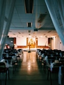 An indoor setting arranged for a formal event, likely a wedding ceremony. Rows of chairs are draped with white fabric, facing a large altar or stage decorated with floral arrangements and a cross. Overhead, the ceiling is adorned with draped fabric and hanging decorations, while the floor reflects a warm light from the stage.