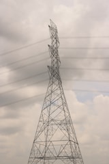 A tall electricity transmission tower stands against a cloudy sky. The structure is composed of a metal lattice framework, supporting several high-voltage power lines that stretch horizontally across the scene. The tower appears to be an essential part of the electrical infrastructure.