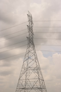A tall electricity transmission tower stands against a cloudy sky. The structure is composed of a metal lattice framework, supporting several high-voltage power lines that stretch horizontally across the scene. The tower appears to be an essential part of the electrical infrastructure.