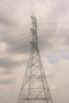 A tall electricity transmission tower stands against a cloudy sky. The structure is composed of a metal lattice framework, supporting several high-voltage power lines that stretch horizontally across the scene. The tower appears to be an essential part of the electrical infrastructure.