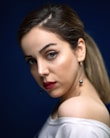 A woman with smooth skin and bold makeup looks over her shoulder. Her eyes feature winged eyeliner, and her lips are painted a deep red. Her hair is pulled back, and she wears a white top over one shoulder, accompanied by dangling pearl earrings. The background is a deep blue, highlighting her features.