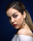 A woman with smooth skin and bold makeup looks over her shoulder. Her eyes feature winged eyeliner, and her lips are painted a deep red. Her hair is pulled back, and she wears a white top over one shoulder, accompanied by dangling pearl earrings. The background is a deep blue, highlighting her features.