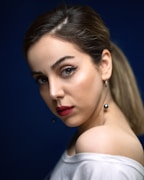 A woman with smooth skin and bold makeup looks over her shoulder. Her eyes feature winged eyeliner, and her lips are painted a deep red. Her hair is pulled back, and she wears a white top over one shoulder, accompanied by dangling pearl earrings. The background is a deep blue, highlighting her features.