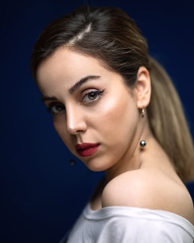 A woman with smooth skin and bold makeup looks over her shoulder. Her eyes feature winged eyeliner, and her lips are painted a deep red. Her hair is pulled back, and she wears a white top over one shoulder, accompanied by dangling pearl earrings. The background is a deep blue, highlighting her features.