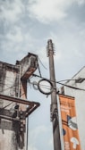 A utility pole with cables and a surveillance camera attached to an old, weathered building façade. The pole also supports a vertical banner with text and simple graphic designs. The sky is cloudy, giving a moody atmosphere.