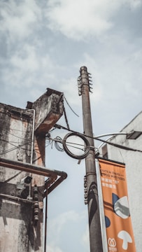 A utility pole with cables and a surveillance camera attached to an old, weathered building façade. The pole also supports a vertical banner with text and simple graphic designs. The sky is cloudy, giving a moody atmosphere.