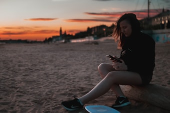 A person sits on a beach during sunset, focused intently on their phone. The sky is a vibrant mix of orange and red hues, and the sandy beach stretches into the distance. A longboard is positioned nearby, hinting at a relaxed and leisurely vibe.