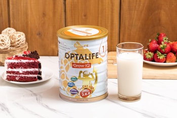 A can of Optalife Grow IQ formula is placed on a marble countertop next to a glass of milk. To the left, there is a slice of red velvet cake on a white plate. In the background, a wooden cutting board holds a pile of fresh strawberries, and a basket contains woven decorative balls. The setting is bright and inviting, with a homely kitchen vibe.