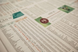 A close-up of a printed financial newspaper or report featuring numbers and text organized in columns and rows. There is a percentage symbol placed within a red circle on a green background, possibly indicating financial data or statistics.